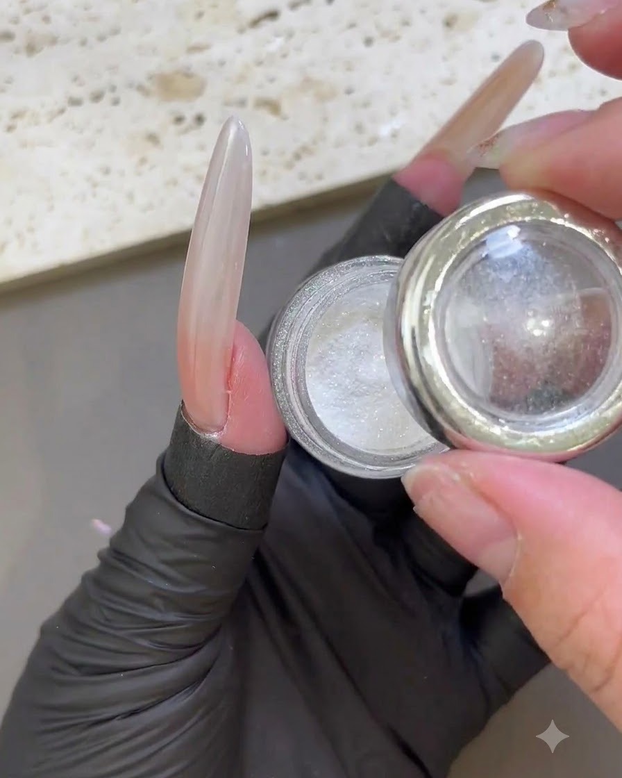 A gloved finger holding an open jar of white iridescent moonlight powder next to a glossy extended nail.