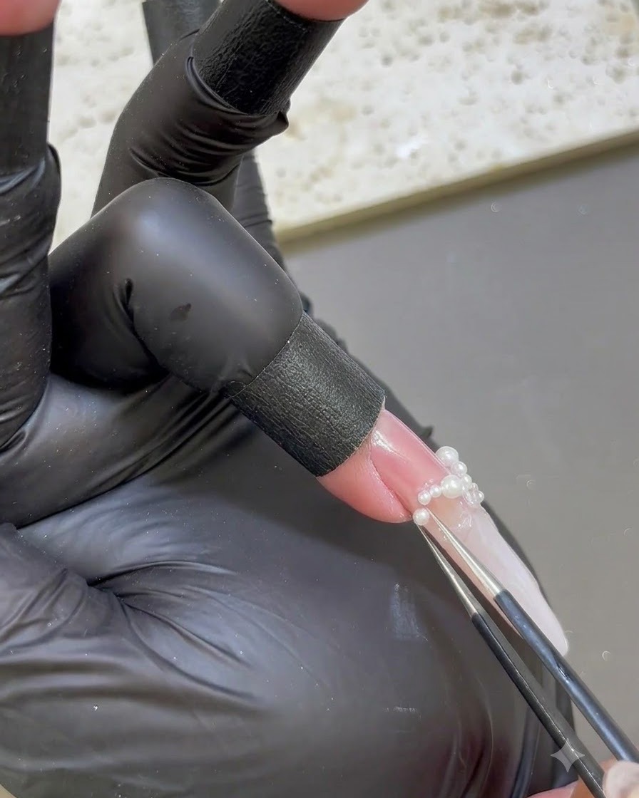 Precision tweezers placing tiny white pearls onto the base of an extended nail.