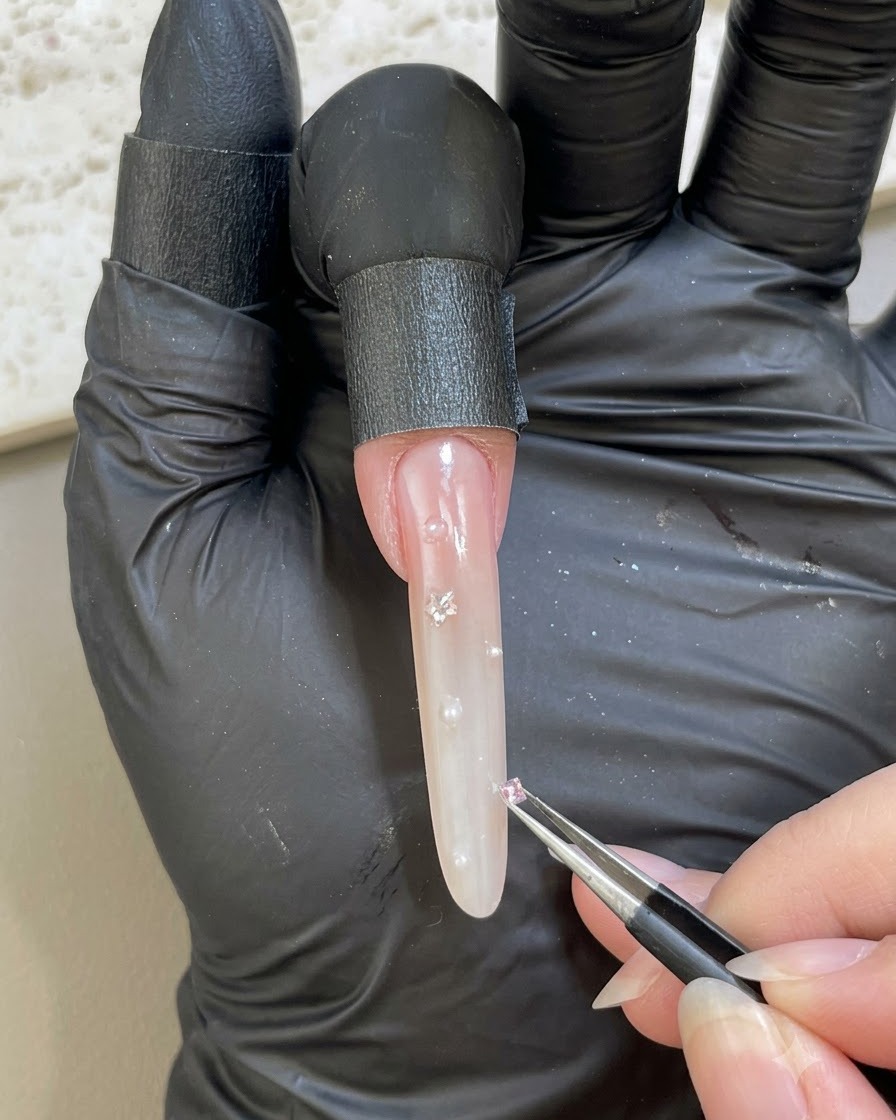 A gloved hand using tweezers to place a small pink rhinestone onto an embellished nail surface.