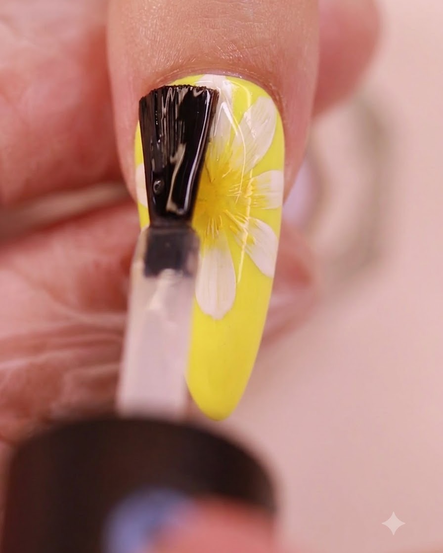 A brush applying a clear layer of no-wipe top coat over the painted daisy nail design.