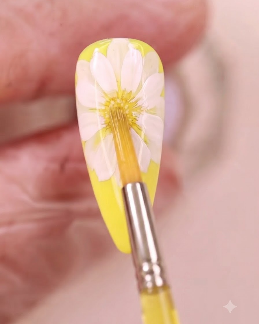 A fine brush applying yellow watercolor liquid to the center of a white daisy nail art design.