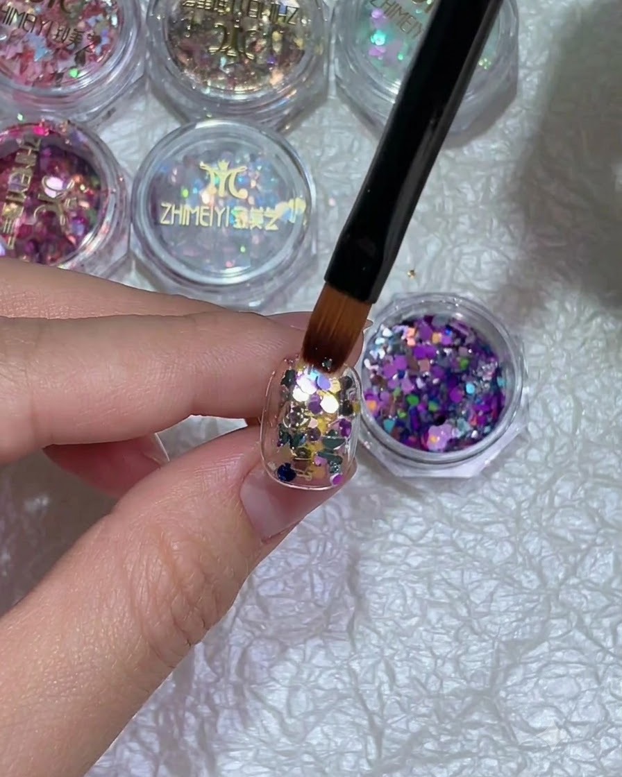 A close-up of a hand with glitter nail art placed under a curing lamp.
