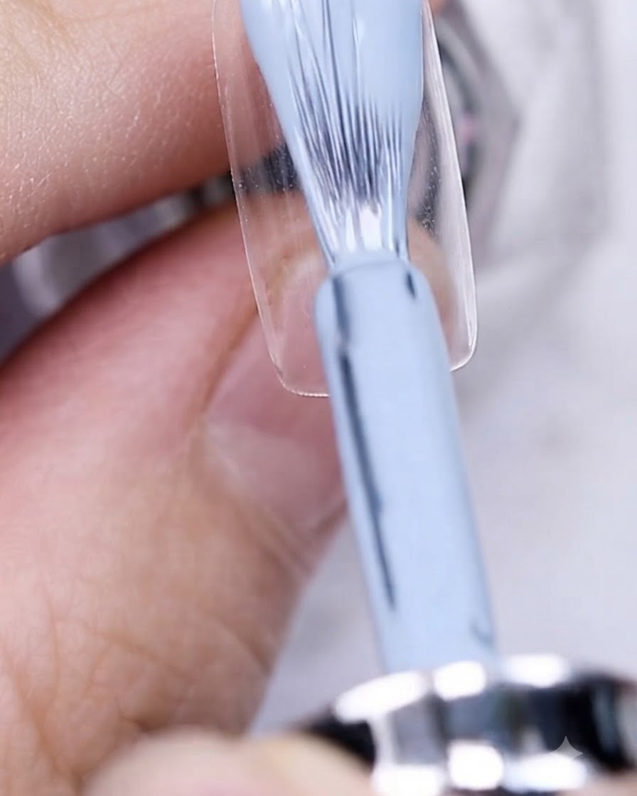 A brush applying grayish-blue gel polish onto a bare nail plate.