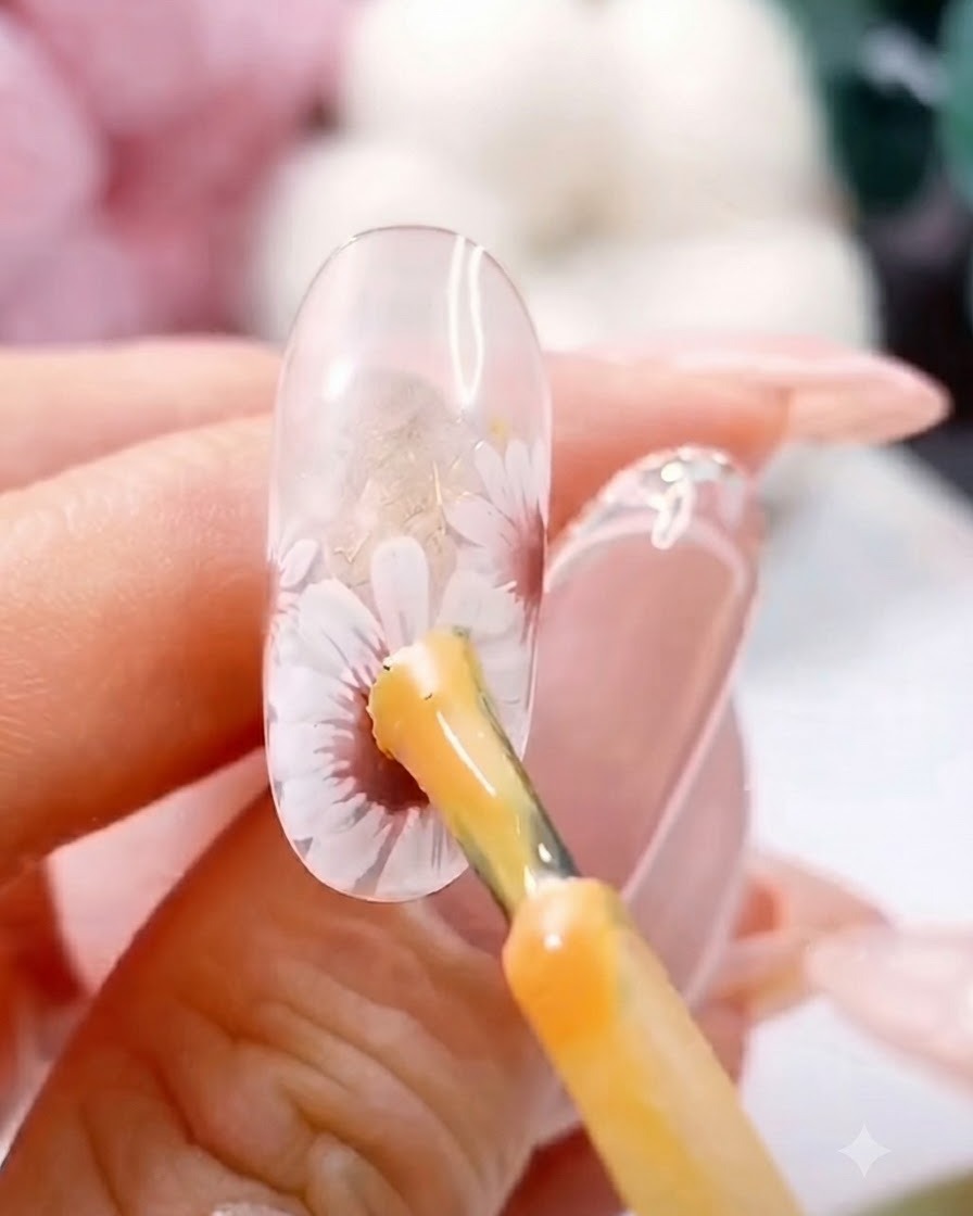 A brush applying a thick yellow gel dot to the center of a painted white daisy on a clear nail.