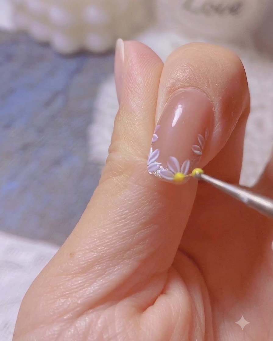 A dotting tool placing a yellow dot at the center of a painted white daisy nail art design.