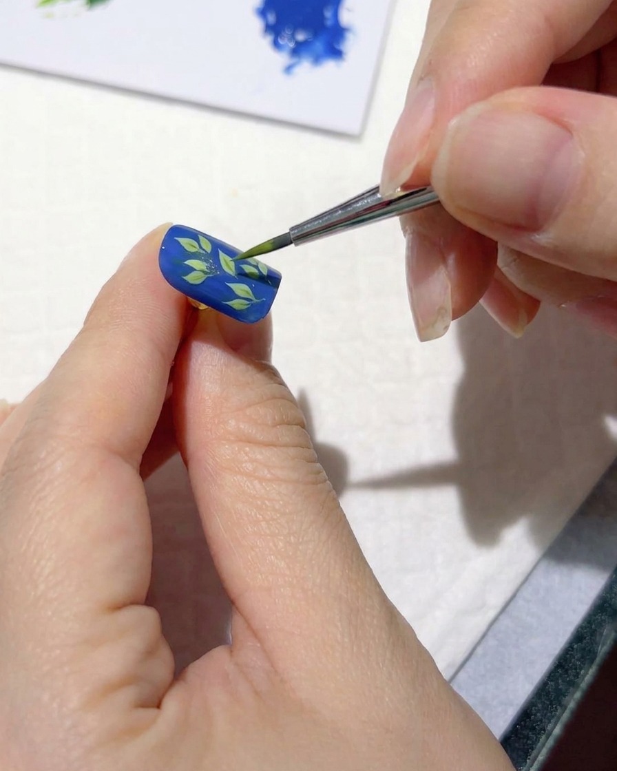 A fine nail art brush adding light green strokes over dark leaves on a blue nail.