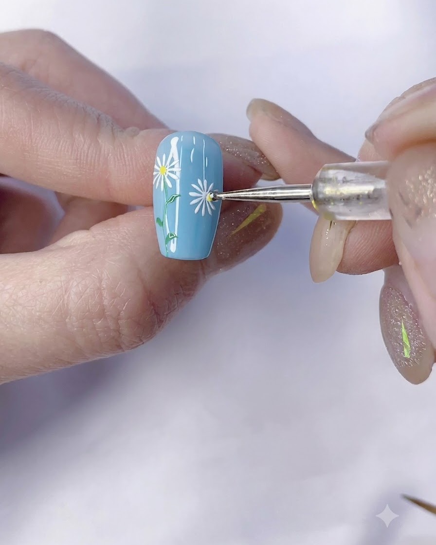 A dotting tool applying a yellow center dot to a white daisy on a blue nail.