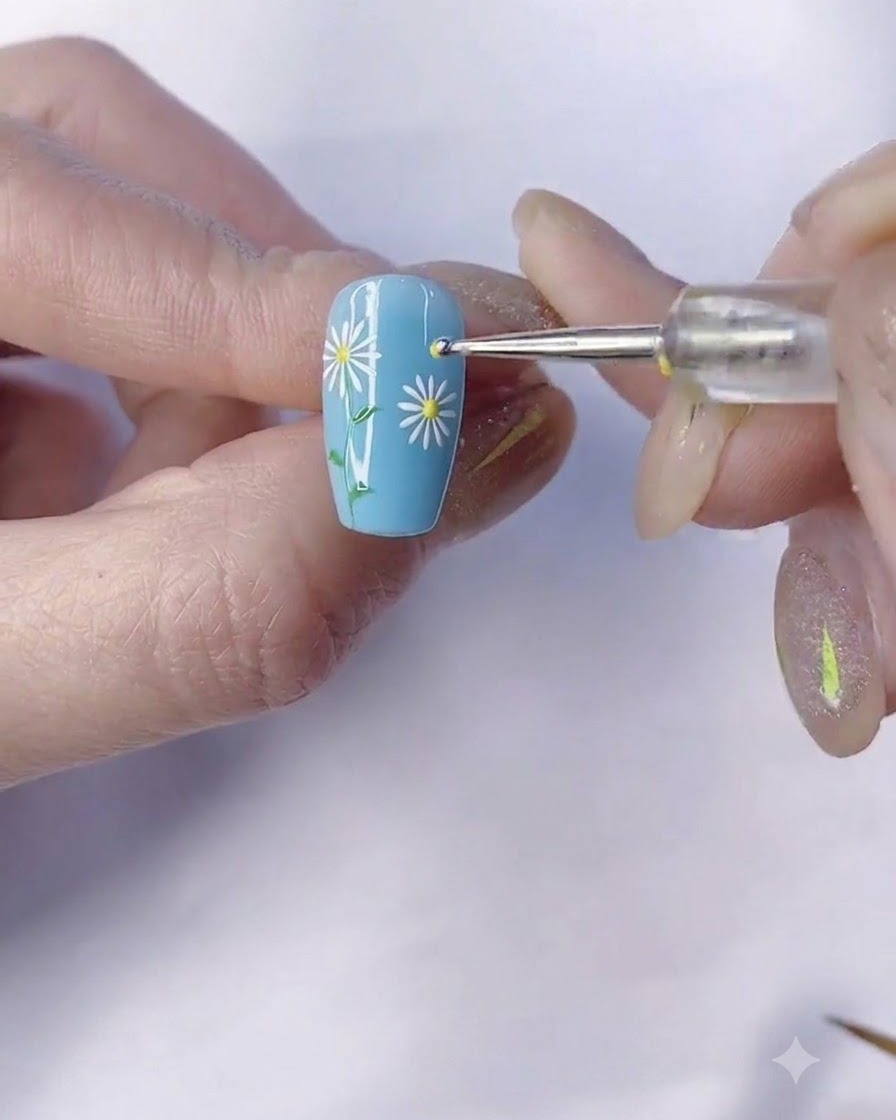 A fine brush drawing a green stem down from the daisy on a blue manicured nail.