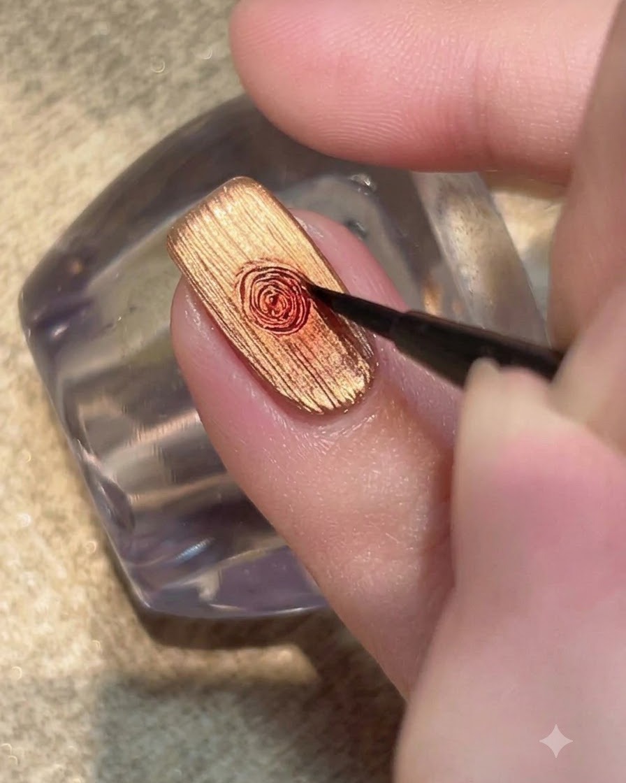A fine detail brush blending a drop of red gel into a circular rose pattern on a metallic gold nail.