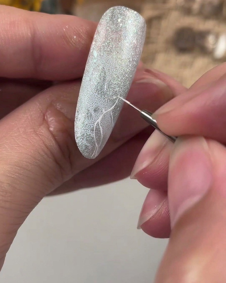 A fine detailing brush drawing a white guideline cross on a textured glitter nail.