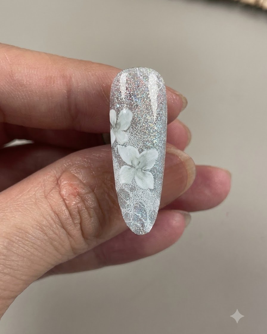 A completed nail showing detailed white and shaded camellia flowers over a sparkling grey-blue lace background.