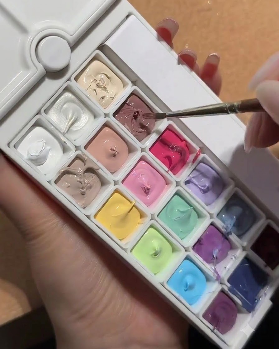A nail brush dipping into a palette containing a square well of white peach nude gel.