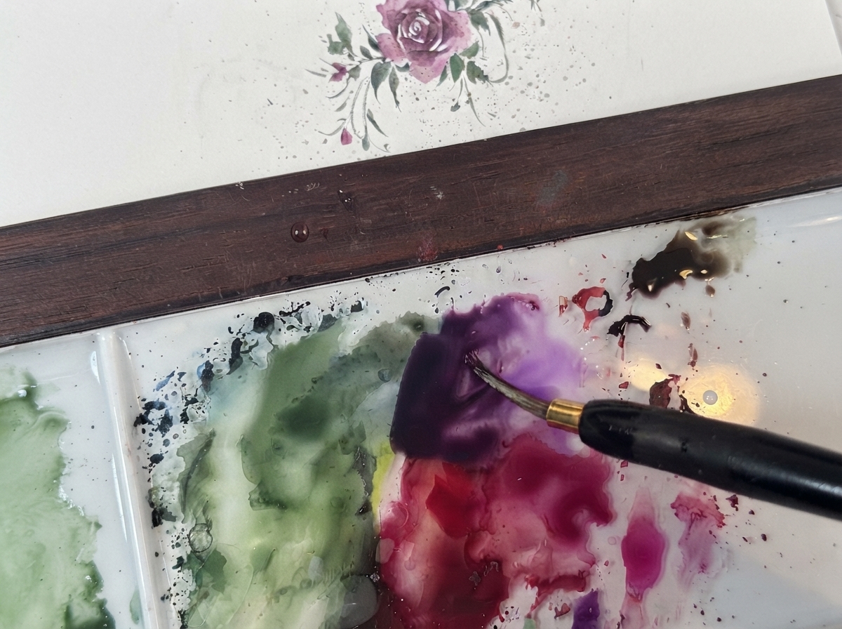 A fine watercolor brush mixing dark purple pigment on a clean white palette next to green washes.