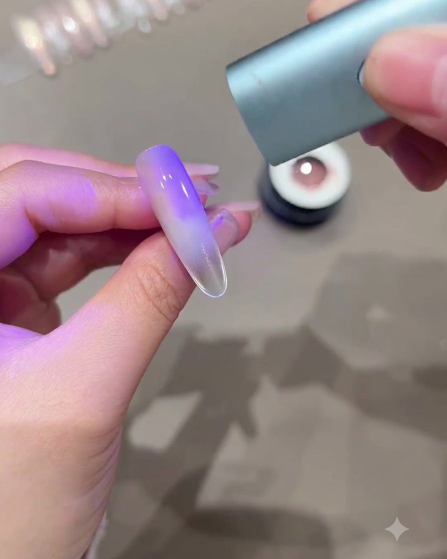 A hand holding a mini UV LED curing light over an elongated nail showing a purple gradient cat-eye effect.