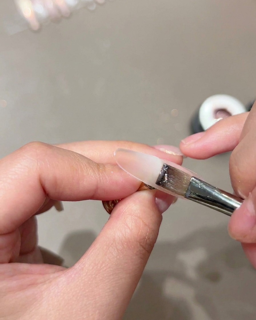 A flat brush applying a sheer nude base layer onto a nail form.