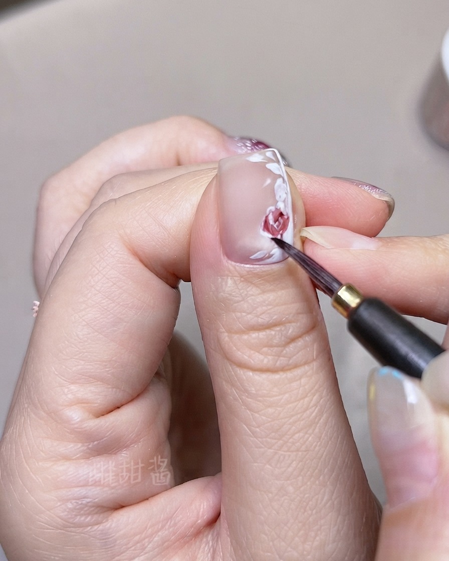 A fine brush layering darker red watercolor tones onto individual painted rose petals and leaves on a fingernail.