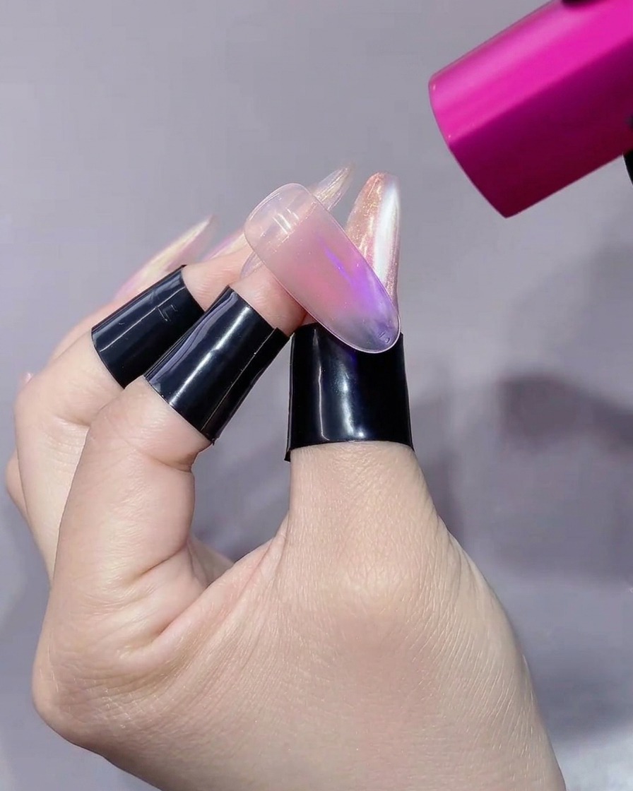 A pink handheld UV/LED lamp pointing directly at a translucent nail extension held on a finger with a black stand.