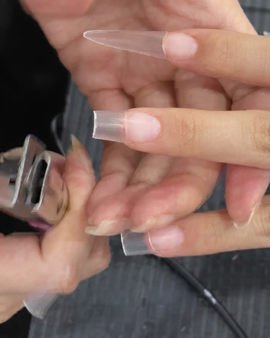 Nail clippers trimming the excess length off a clear artificial nail extension.