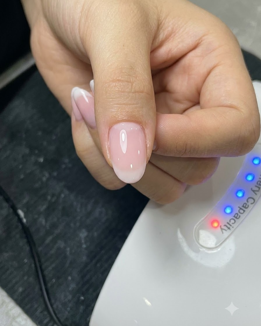 A brush applying a top coat layer onto a manicured nail in front of a UV lamp.