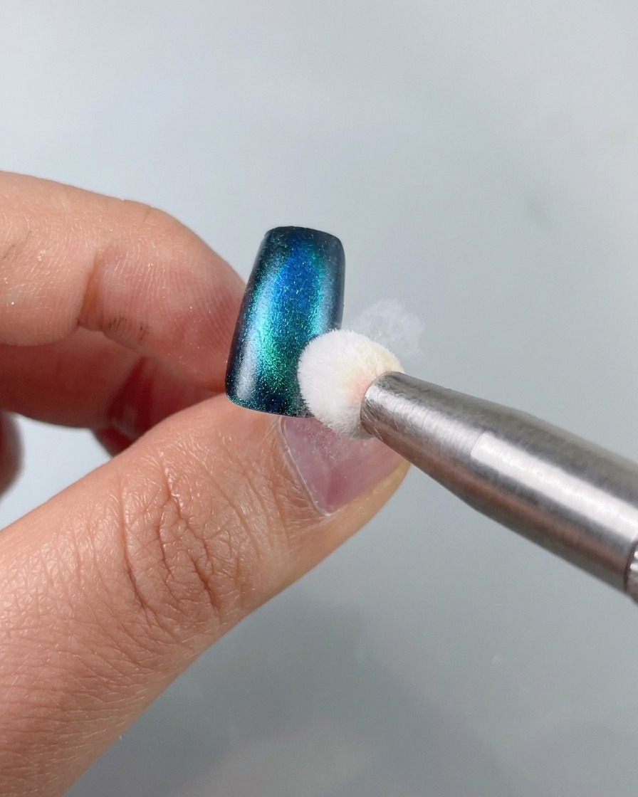 A sponge tool dabbing black stamping gel onto the edge of a blue-green cat-eye nail piece.