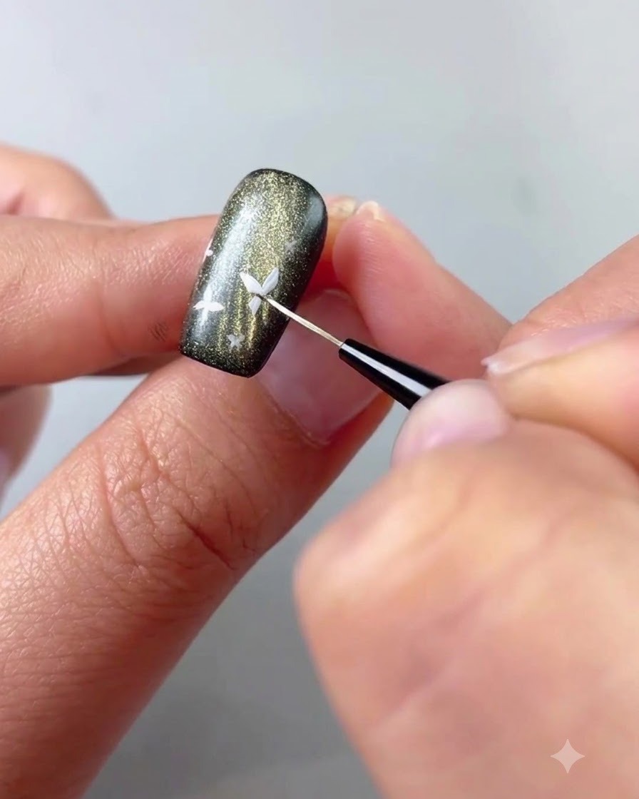 A close-up of a fine liner brush applying white gel to paint a small butterfly detail on a dark cat-eye nail.