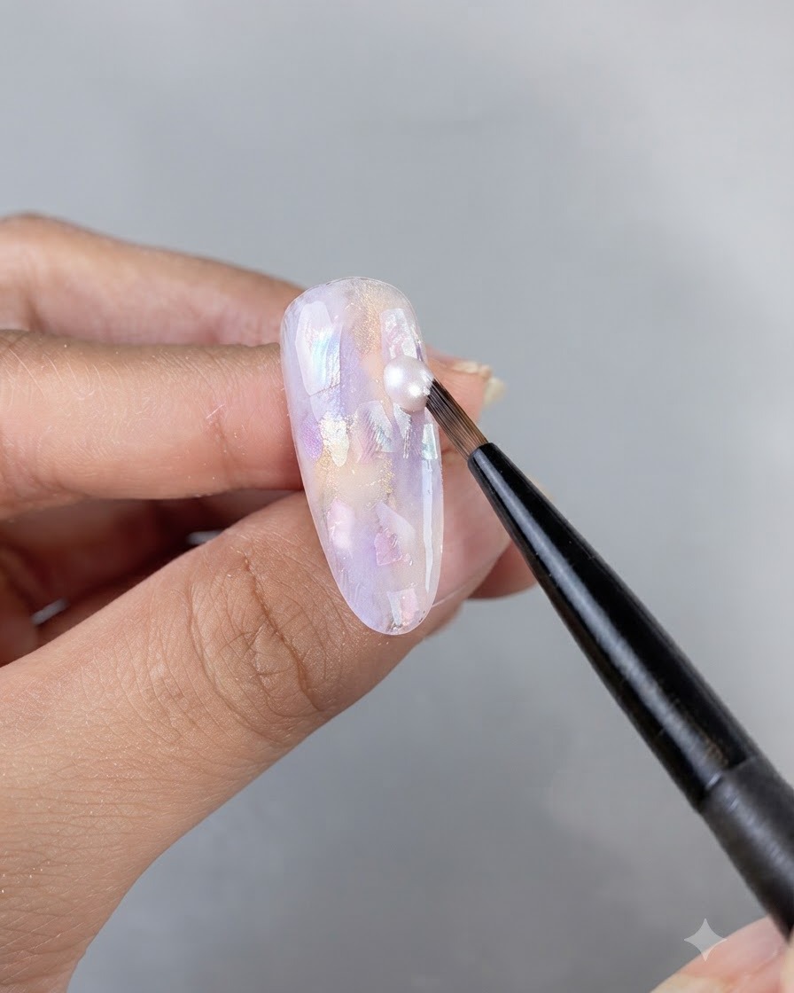 A fine art brush applying a drop of shimmering gel onto an ornate, shell-embedded nail surface.
