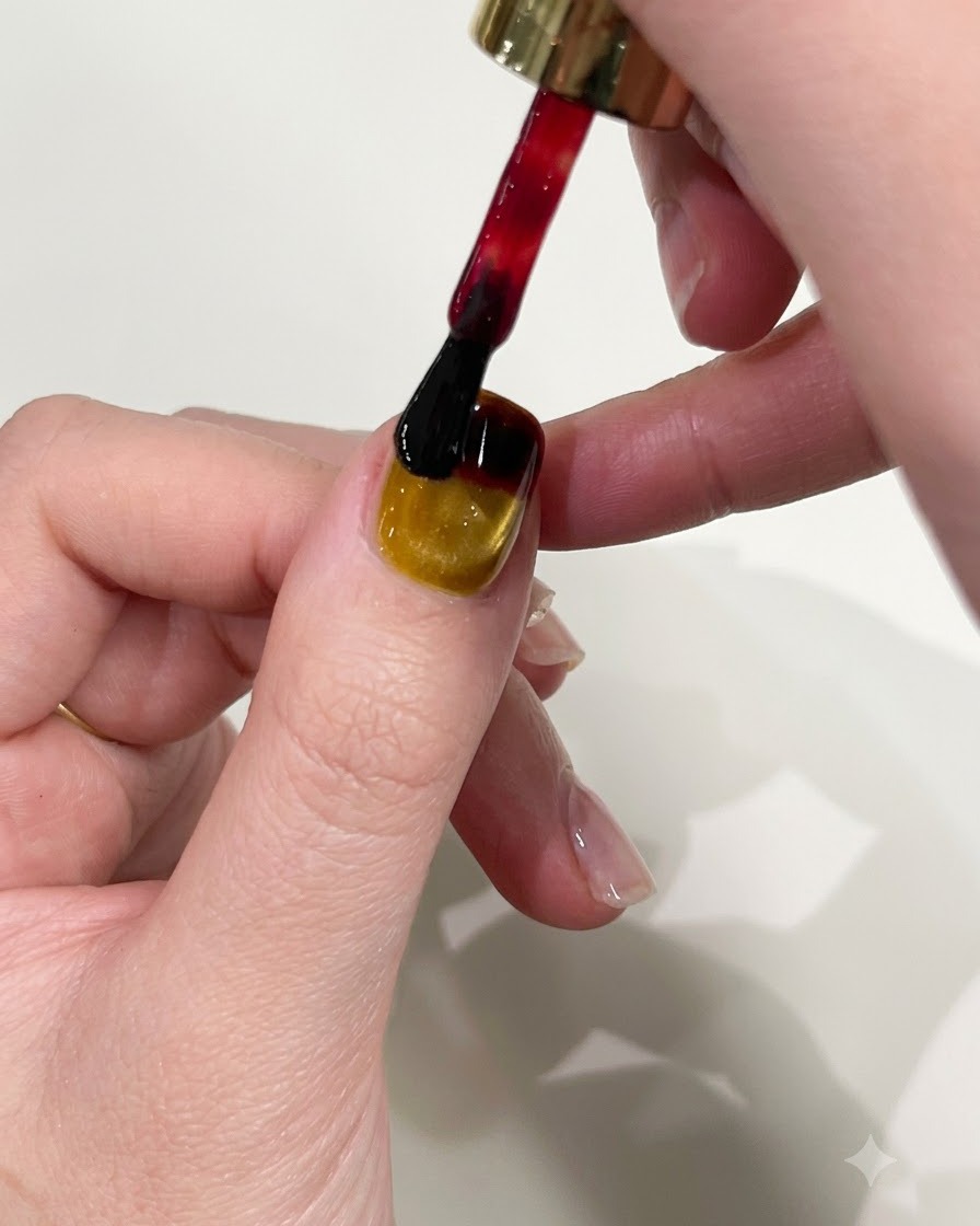 A nail polish brush applying red gel to the upper edge of a yellow cat-eye manicured nail.