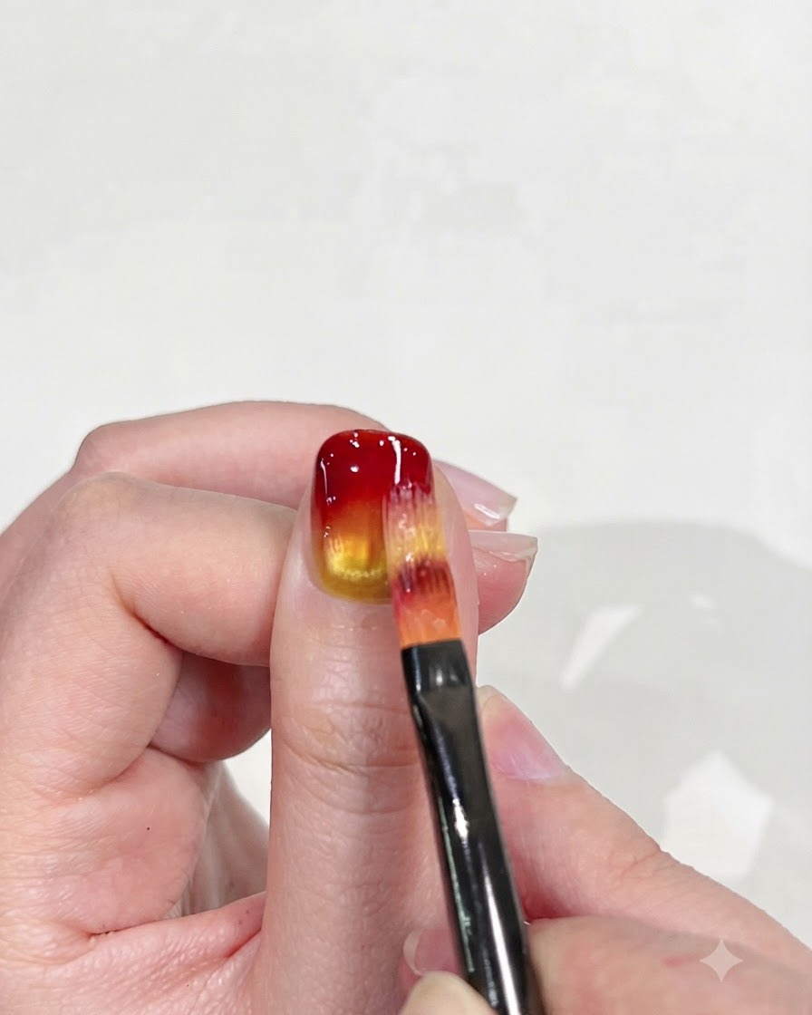 A flat nail brush carefully blending the red tip downward into the yellow base to refine the gradient effect.