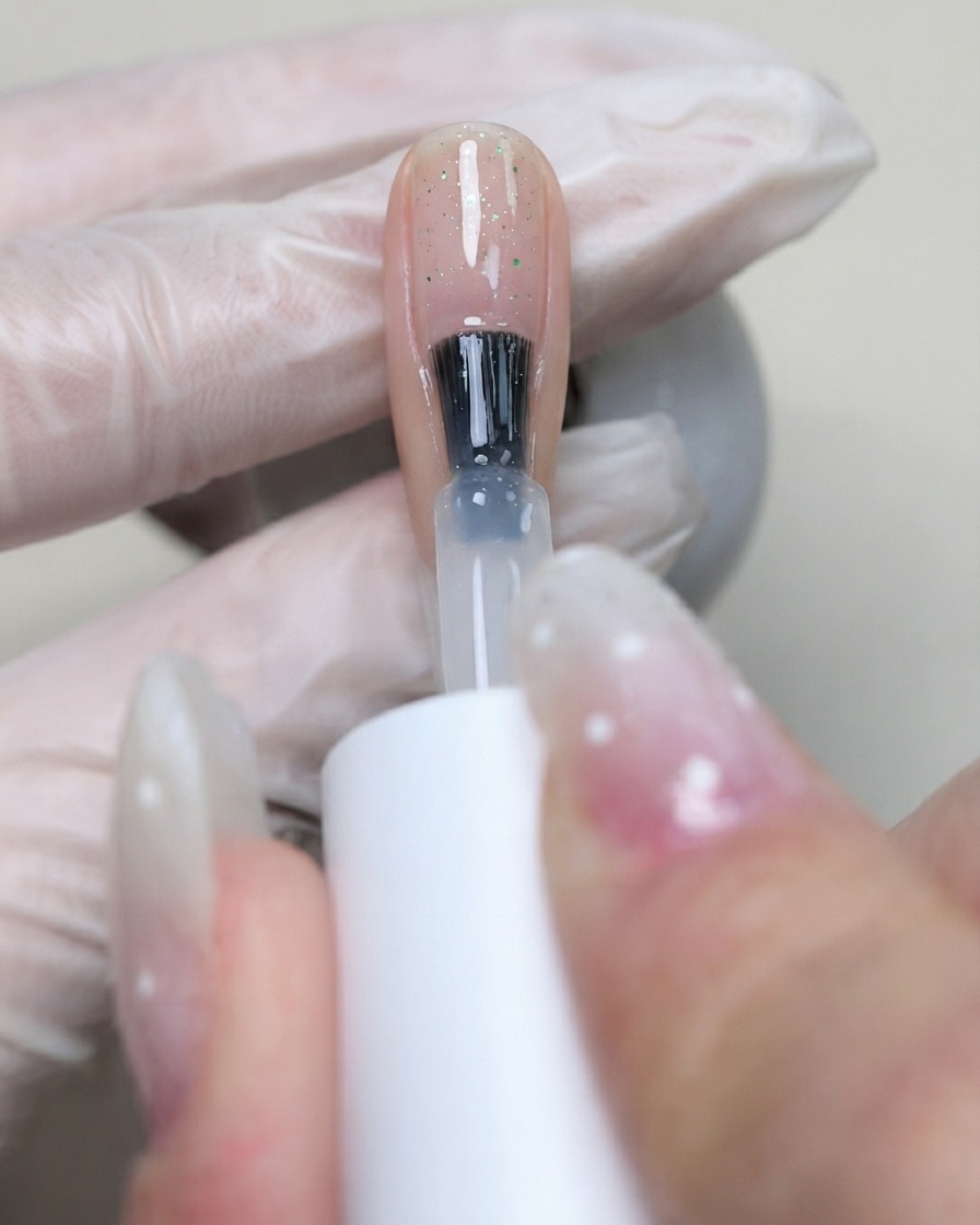 A brush applying clear C7 flexible base gel over the nail surface to lock in the placed sequins.