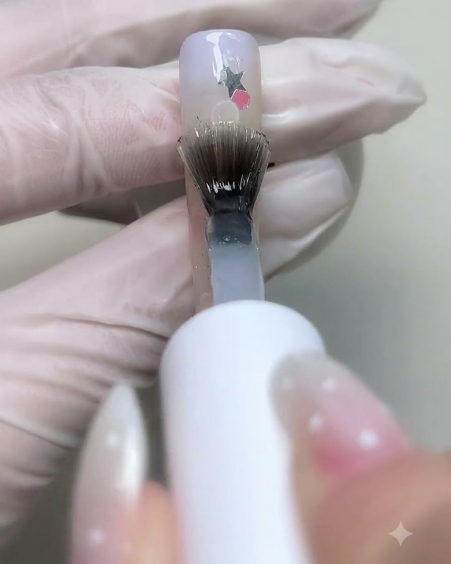 A brush applying clear base gel over decorated nails with star sequins and bubbles.