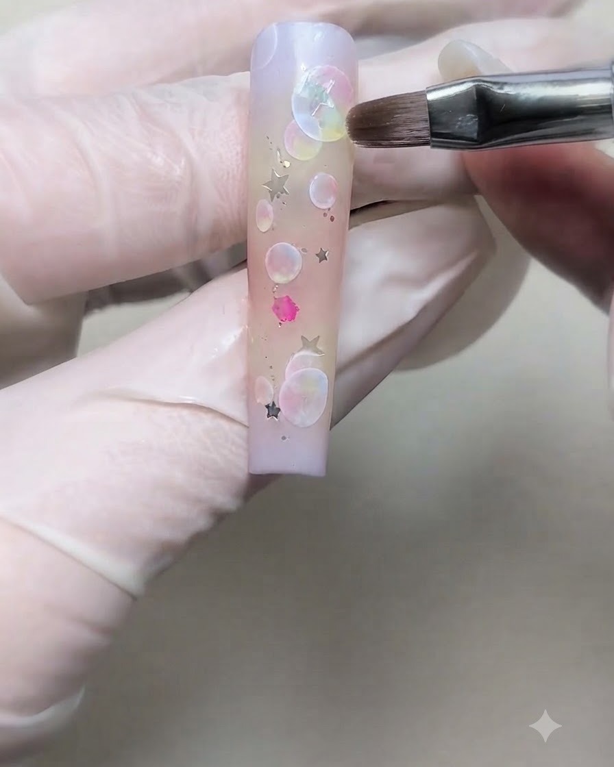 A gloved hand placing a silver star sequin onto a gradient nail with a fine brush.