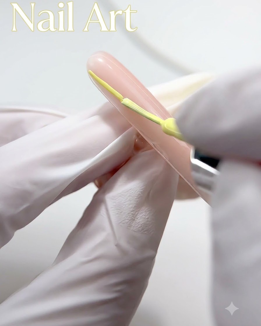 A gloved hand using a fine brush to apply yellow gel polish along the tip of a nude-colored nail.