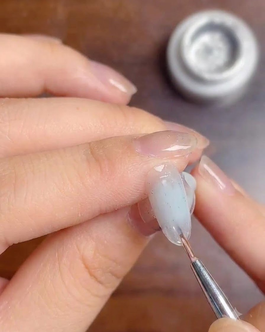 A fine brush applying a layer of clear top coat gel over silver glitter lines on a milky white gradient nail.