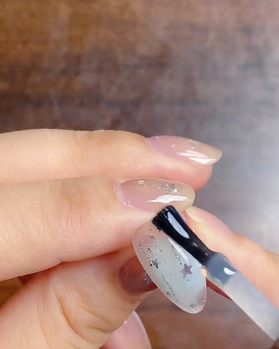 A nail brush applying a final glossy top coat over a milky gradient nail decorated with silver glitter and star stickers.