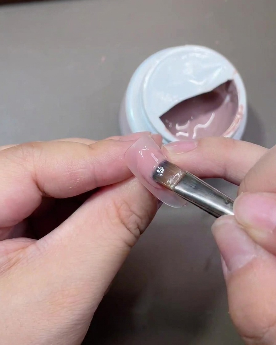 A gel brush applying a smooth coat of translucent nude polish onto a nail surface.