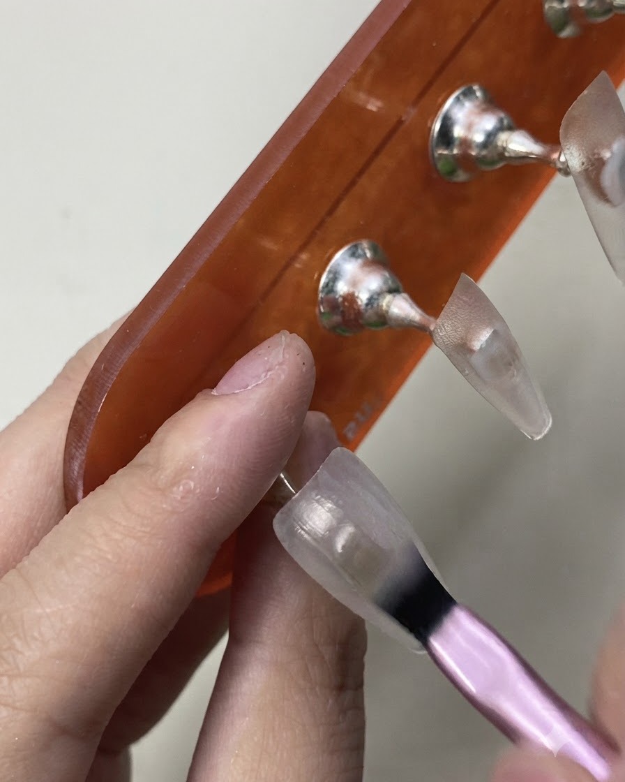 A brush carefully applying the initial coat of sheer milky white gel polish onto a practice nail tip.