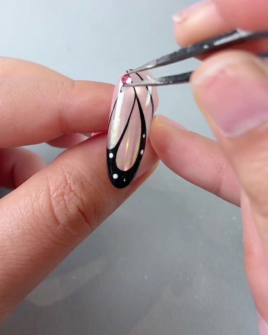 A pair of tweezers placing a pink crystal rhinestone onto the top of a butterfly wing nail art design.