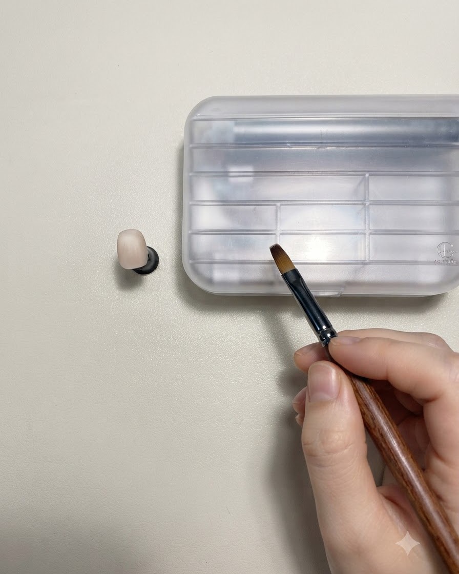 A hand holding a wooden-handled nail art brush next to a nude-colored practice nail on a stand.
