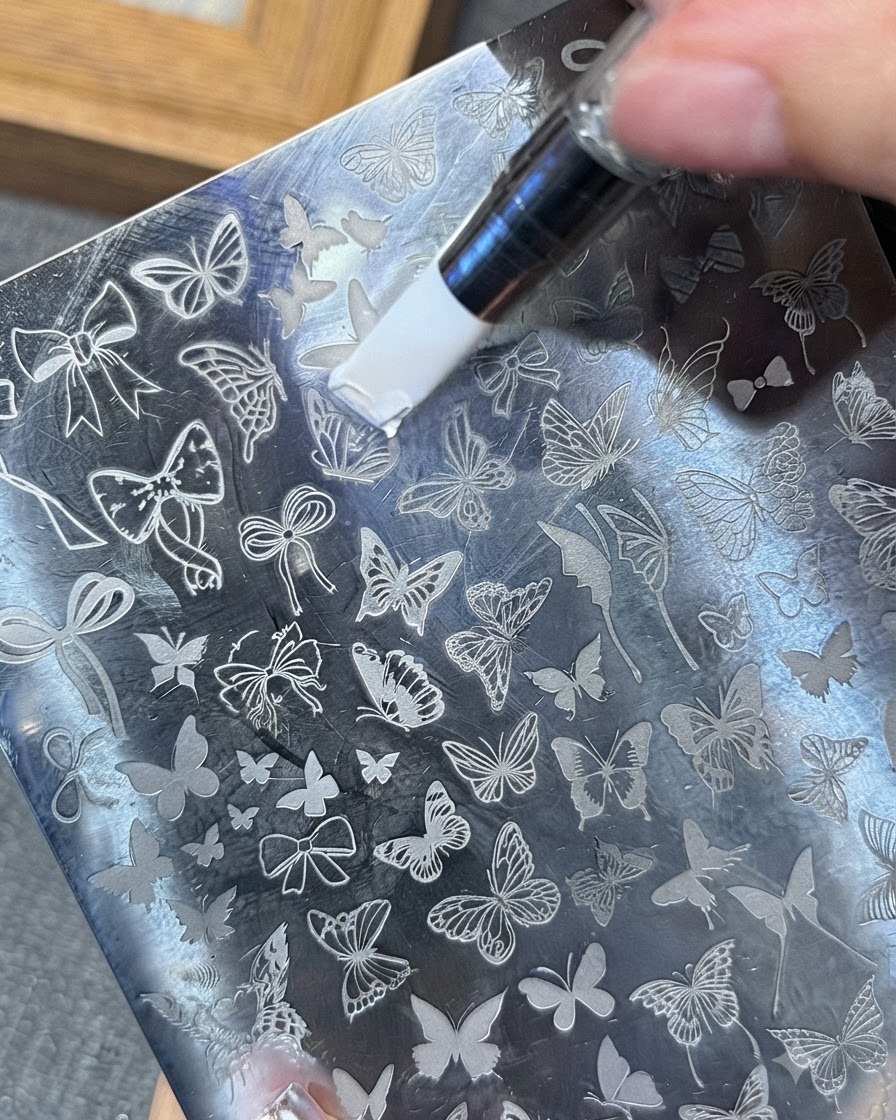A tool spreading white stamping gel across a butterfly pattern on a metal stamping plate.