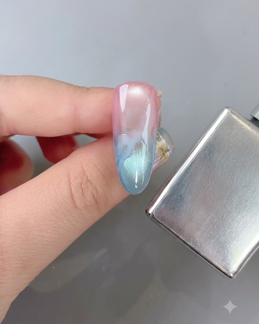 A nail brush softly blurring the edges of a white cloud pattern on a multi-colored gradient nail next to a magnetic stick.