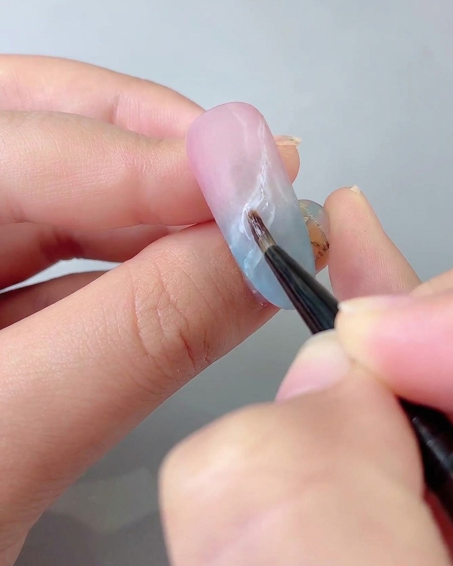 A fine detail brush drawing delicate ribbon lines on the nail surface with sticky gem glue.