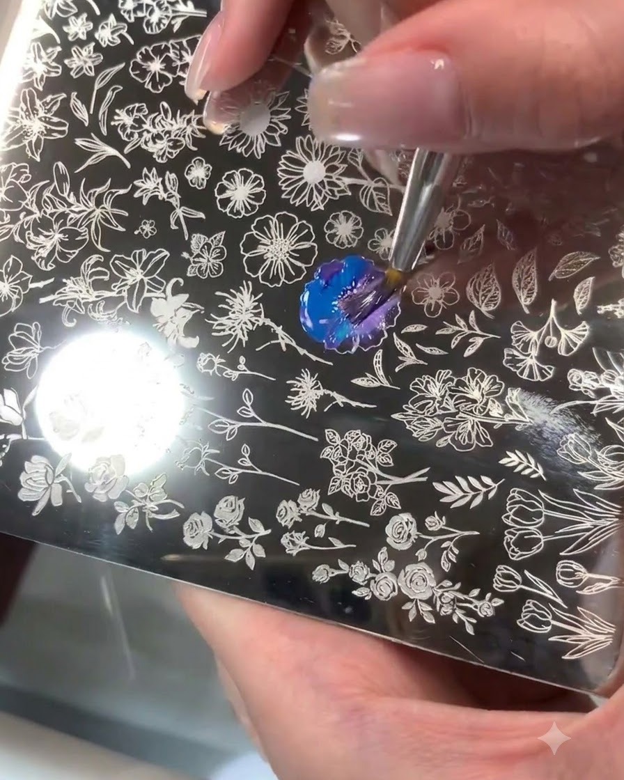 A steel stamping plate with an engraved floral pattern where blue and purple gel is being mixed with a brush.