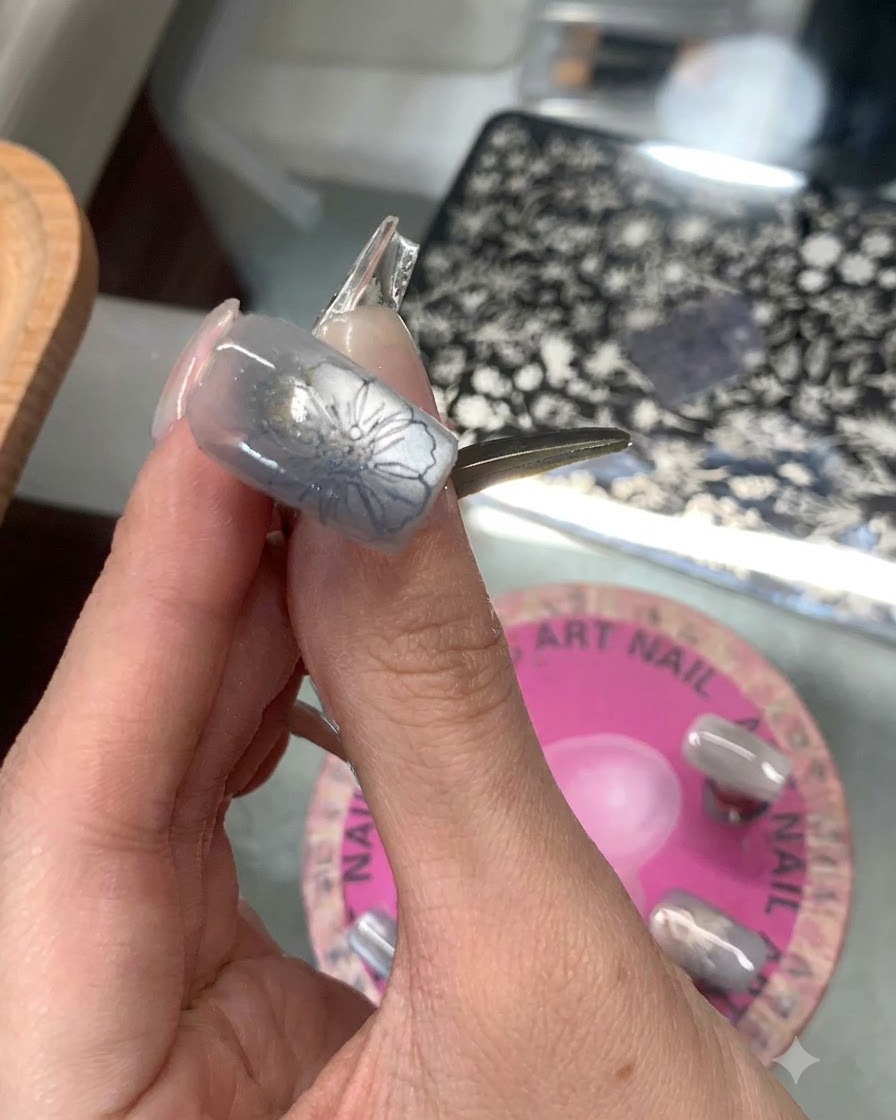 A clear silicone stamper pressing a blue floral design onto the tip of a greyish-blue manicured thumb nail.