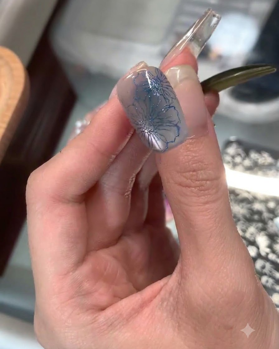 A clear silicone stamper applying delicate blue floral stamping onto the tip of an index fingernail.