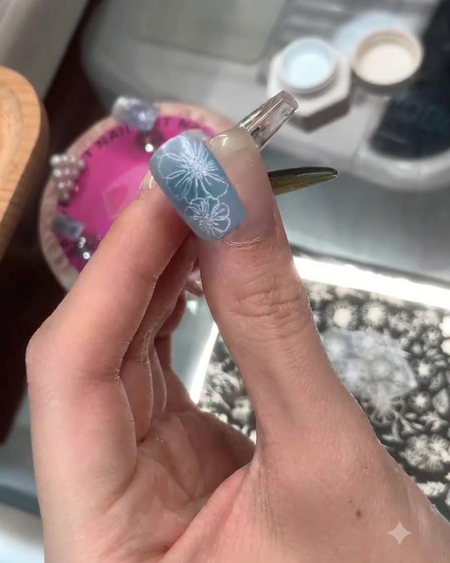 A manicured finger displaying a completed blue base with crisp white stamped floral details on the nail tip.