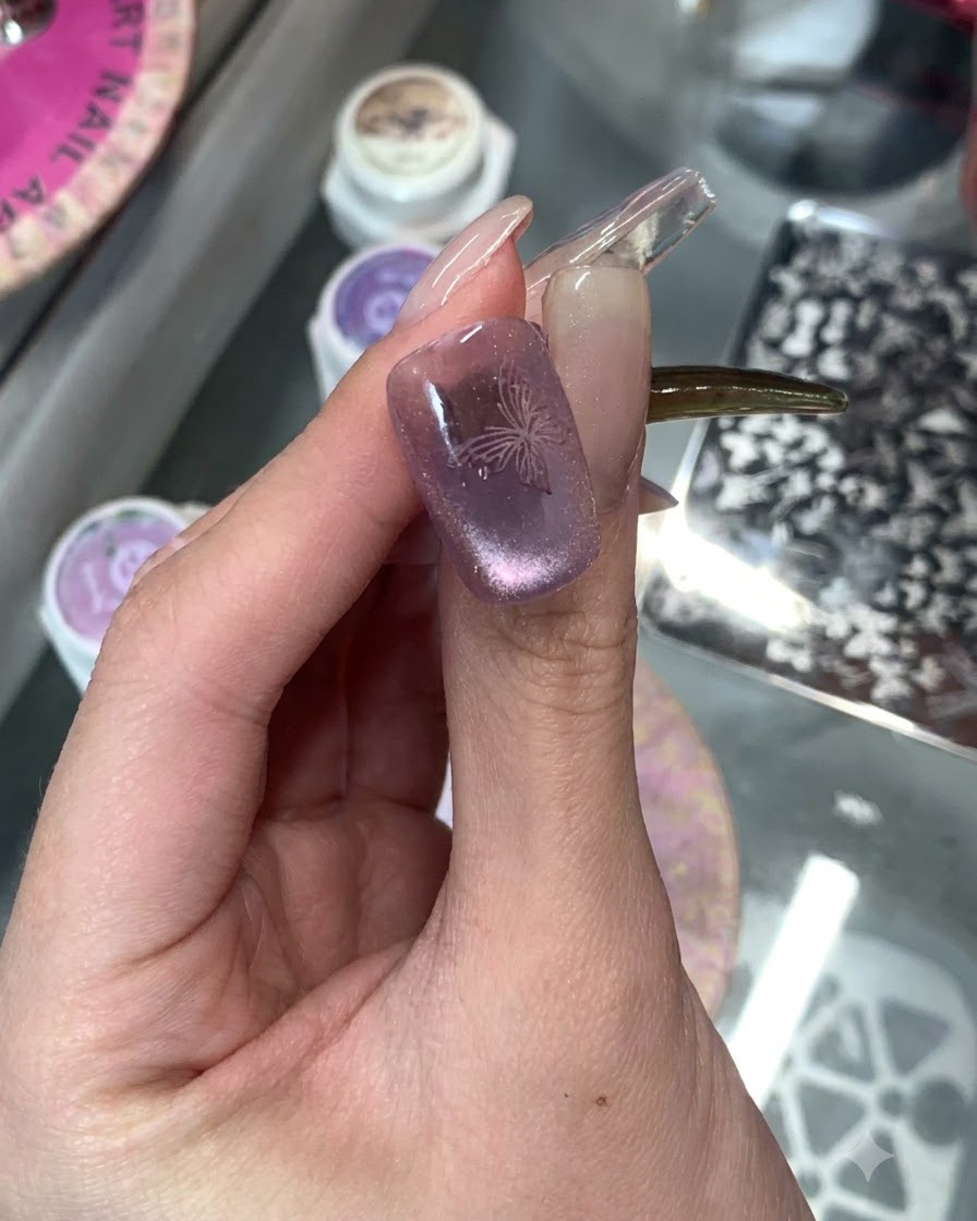 A close-up of a purple cat-eye manicured nail with a delicate stamped butterfly design on top.