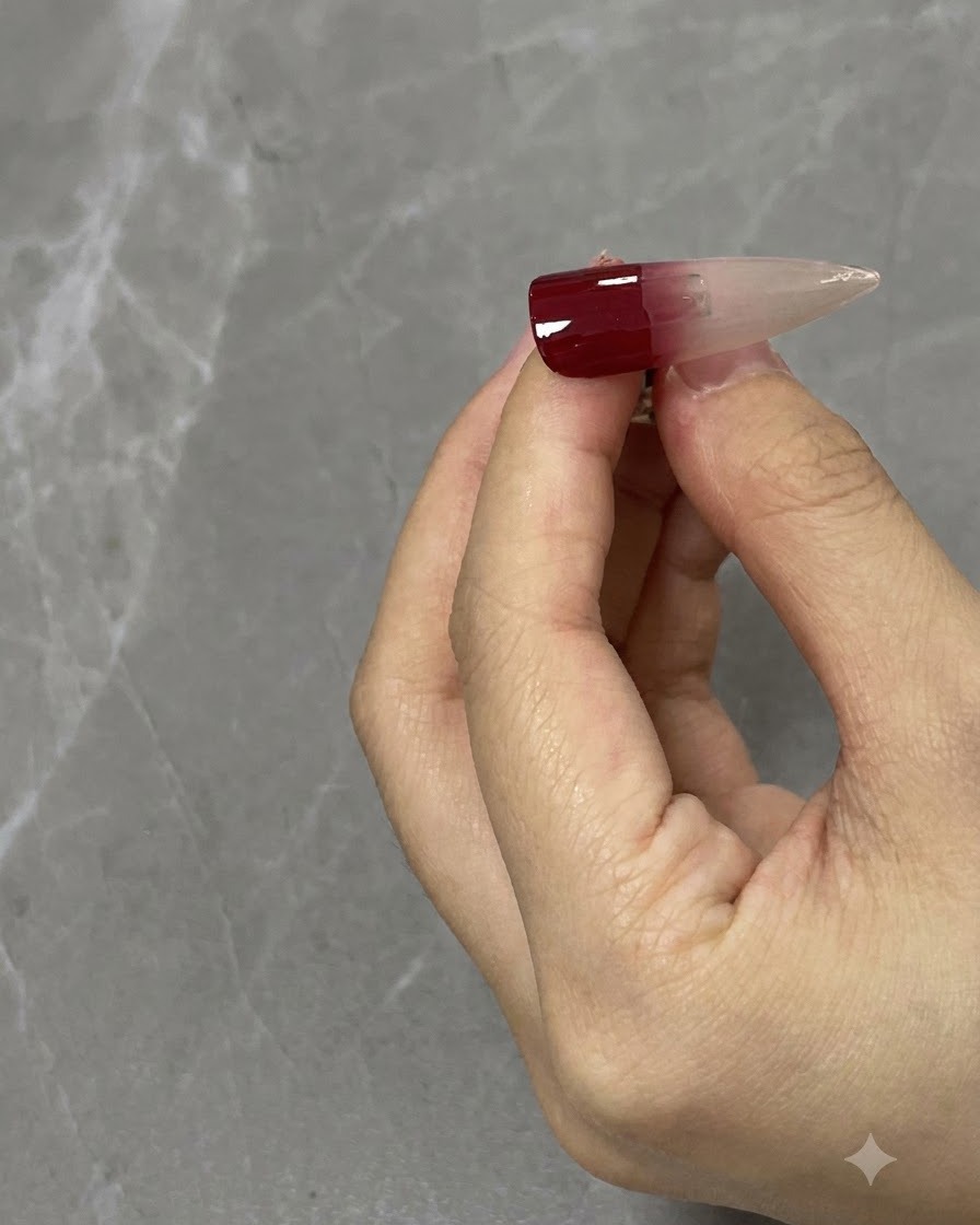 A close-up of a nail brush blending a wine red gradient downwards on a neutral nail tip.