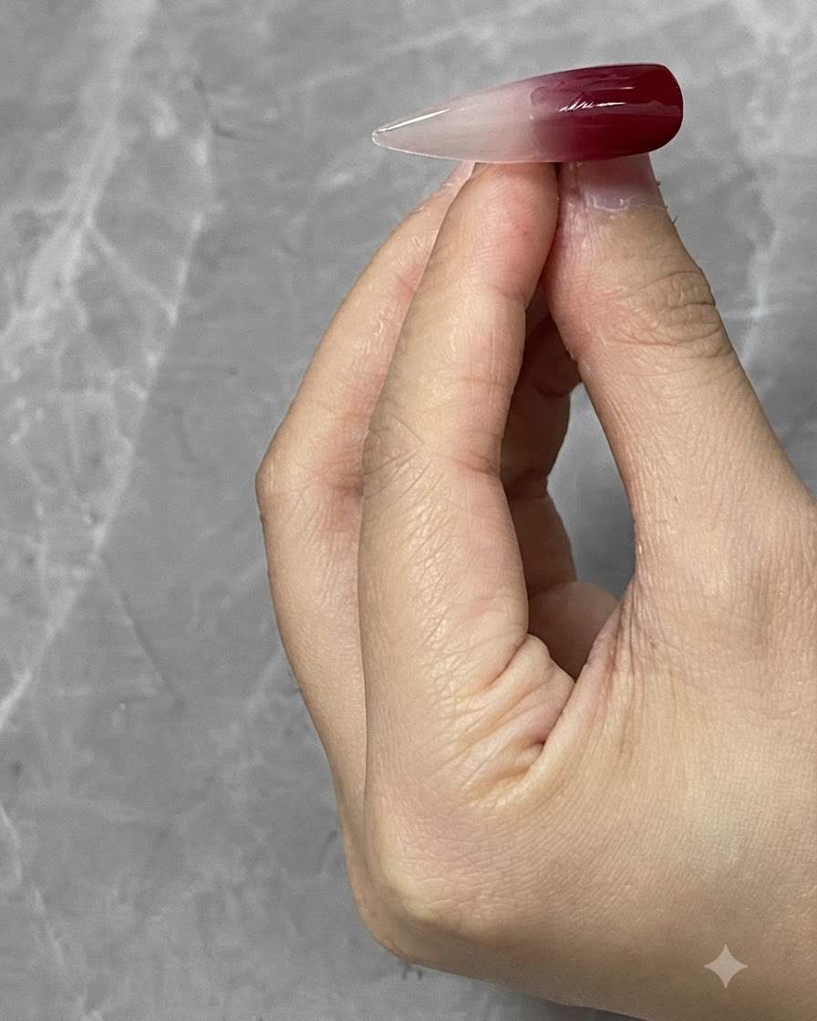 A close-up of a nail tip held by a hand, showing a rich wine red solid color blending into a translucent gradient.