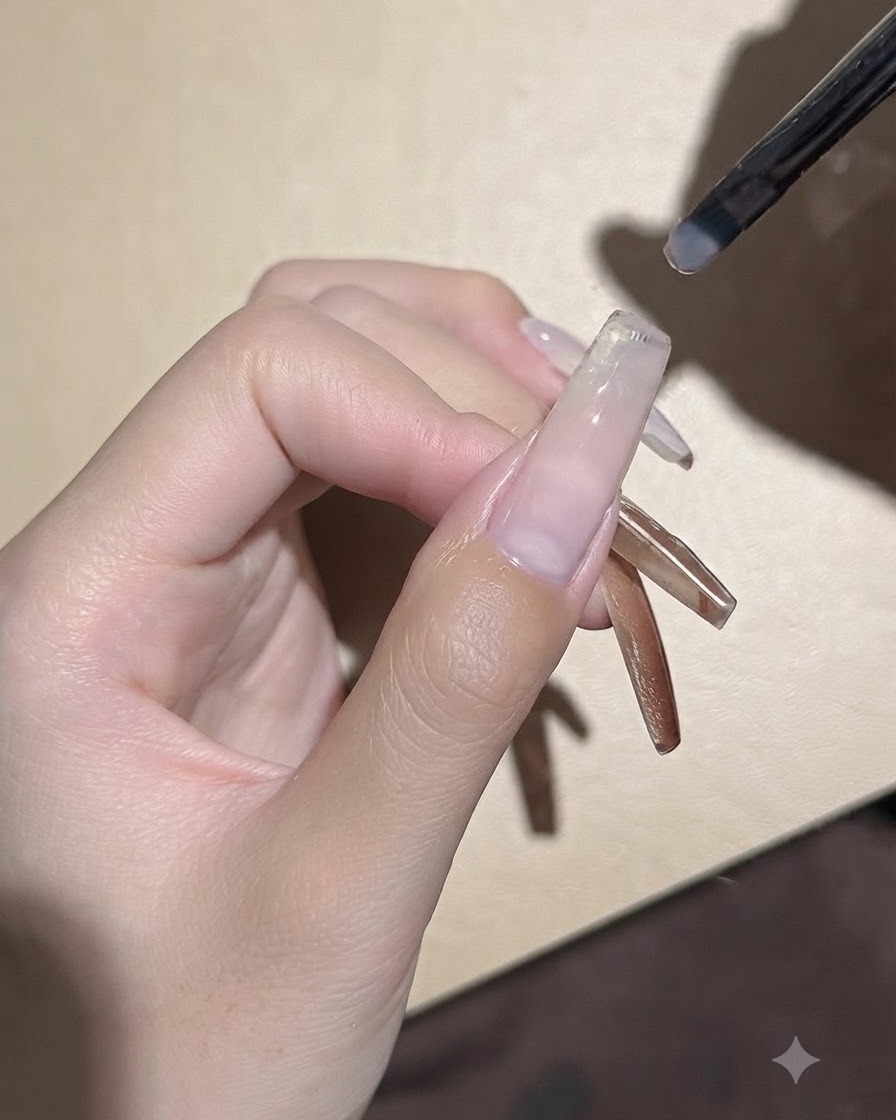 A small gel brush applying a smooth coat of clear base gel onto an extended nail plate.