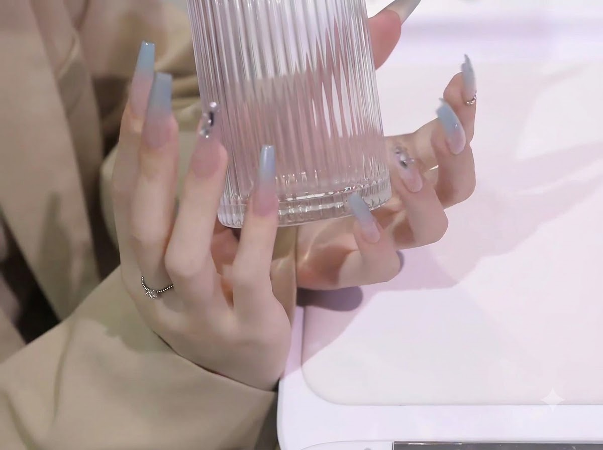 Both hands lifting a clear ribbed glass container, displaying the length and curvature of blue gradient nail extensions.